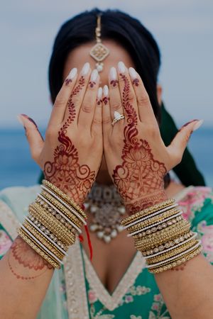 Intricate henna designs adorn hands with gold bangles against an ocean backdrop.