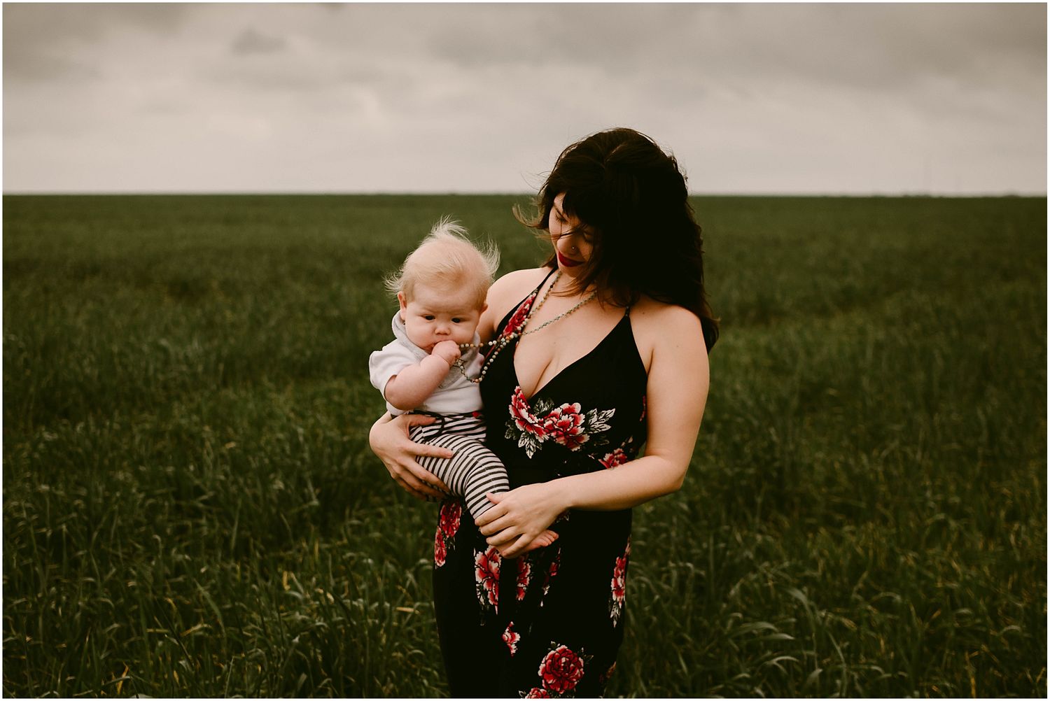 Figure in black floral dress holds baby against dramatic cloudy sky in grassy field.
