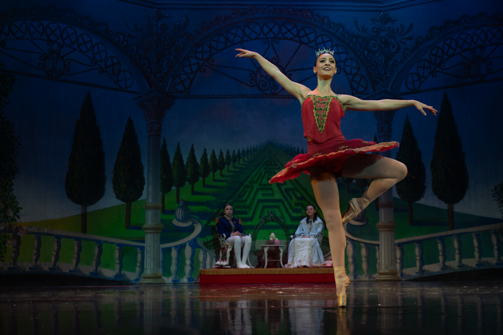 Ballet dancer in red tutu performs graceful leap against painted garden backdrop.