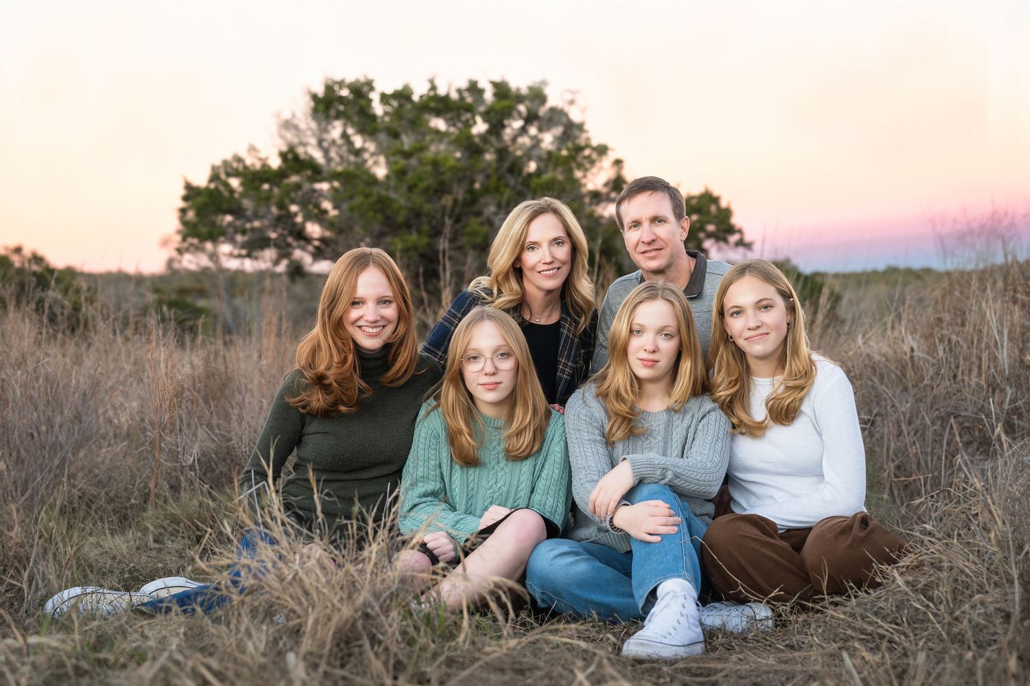 mom, dad and four teen daughers sit in field at Phil Hardberger Park with pinks and purples in sky