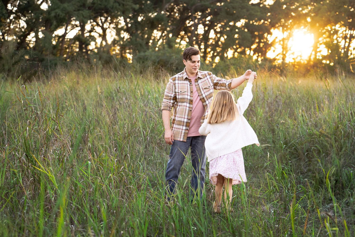 dad wearing a plaid shirt twirling his daughter in a pink printed dress in a field with the sun setting in Boerne, TX