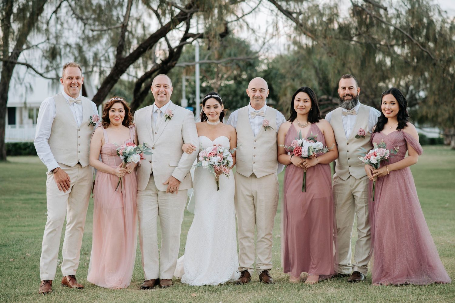 Wedding party in pink and beige attire poses together on a grassy lawn under trees.