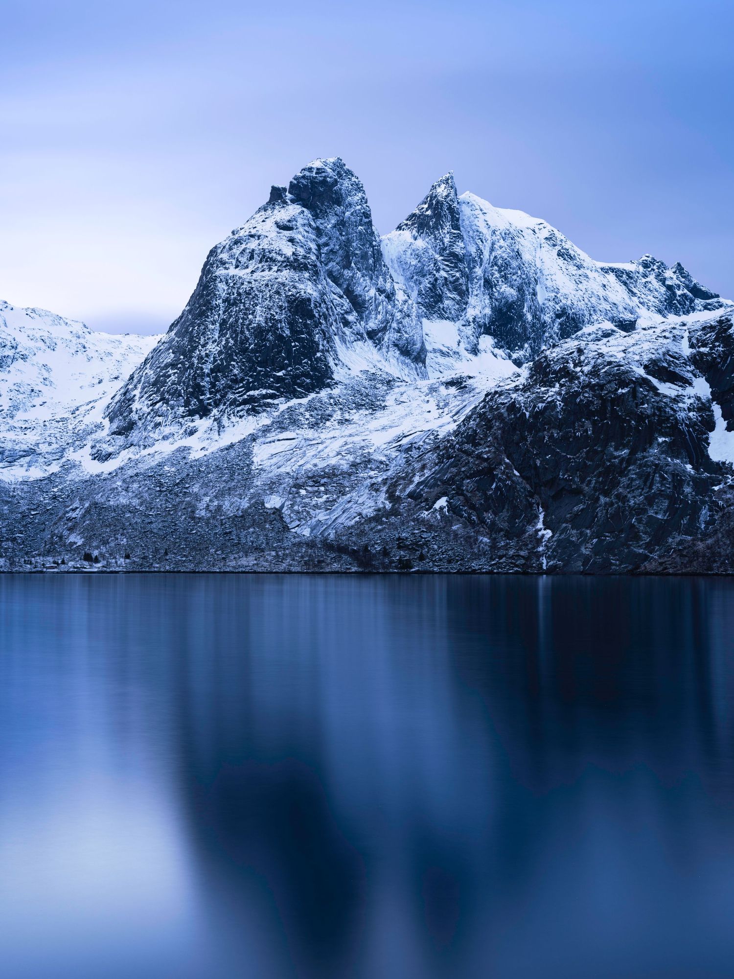 Majestic snow-covered mountains tower over calm blue waters in a serene winter fjord landscape.