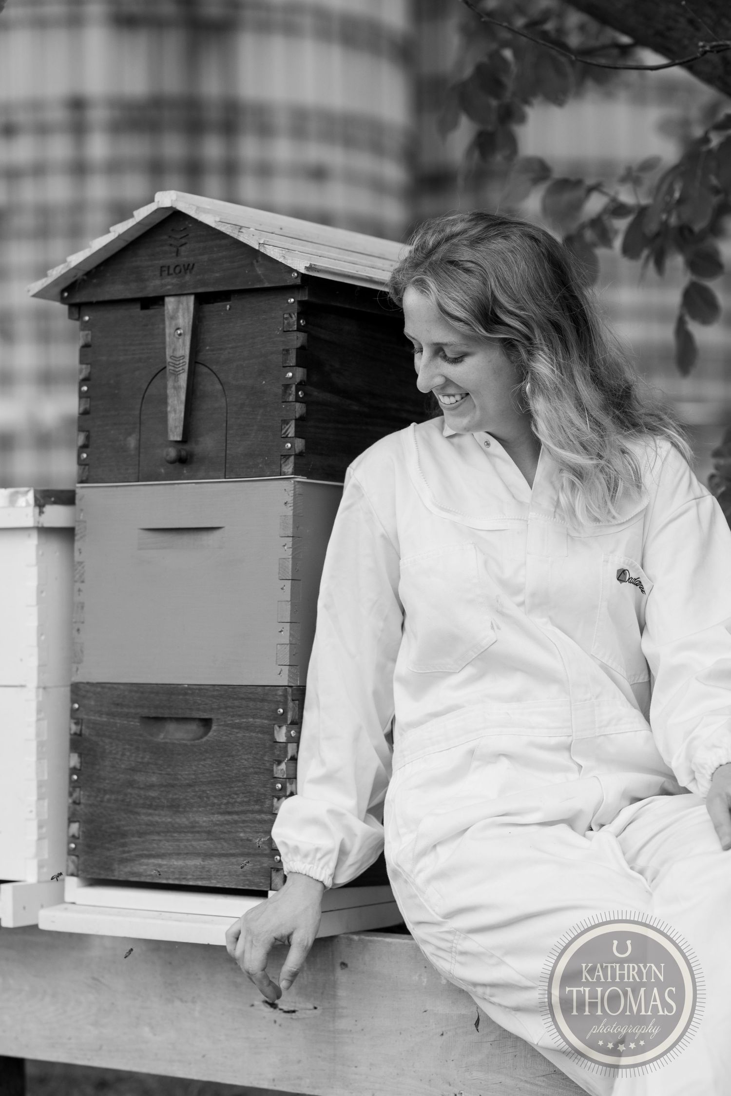 Black and white image of a beekeeper sitting beside wooden hive boxes under tree branches.