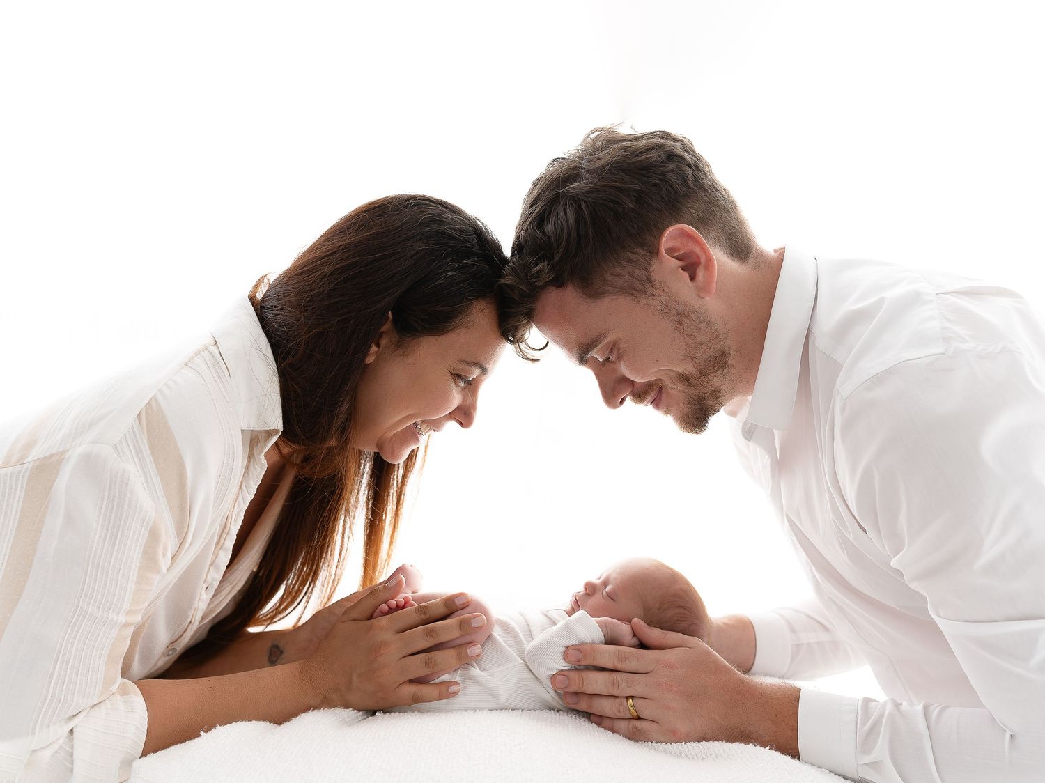 A couple in white clothing leans in close together while looking down with joy.