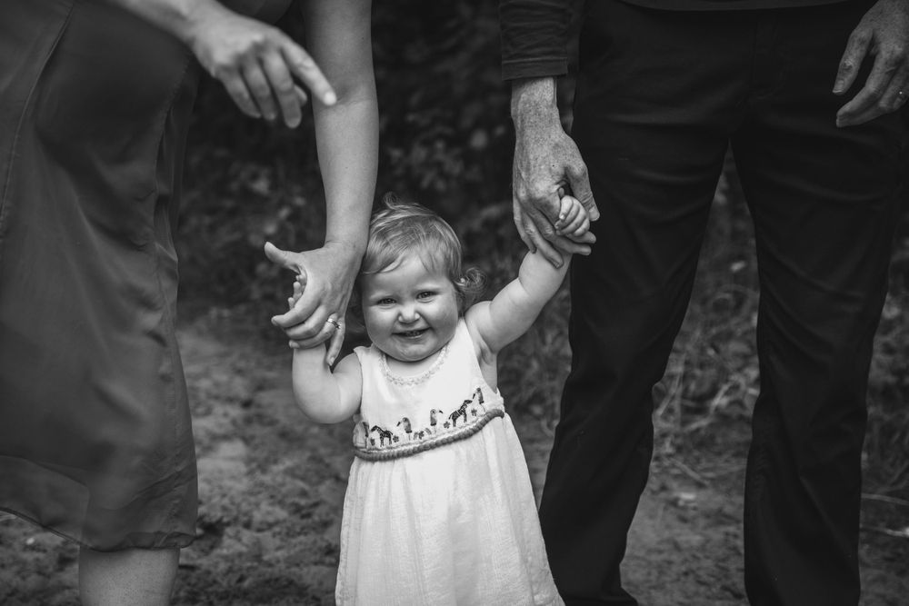 A black and white photo of a toddler in a dress being held by adults' hands while walking.