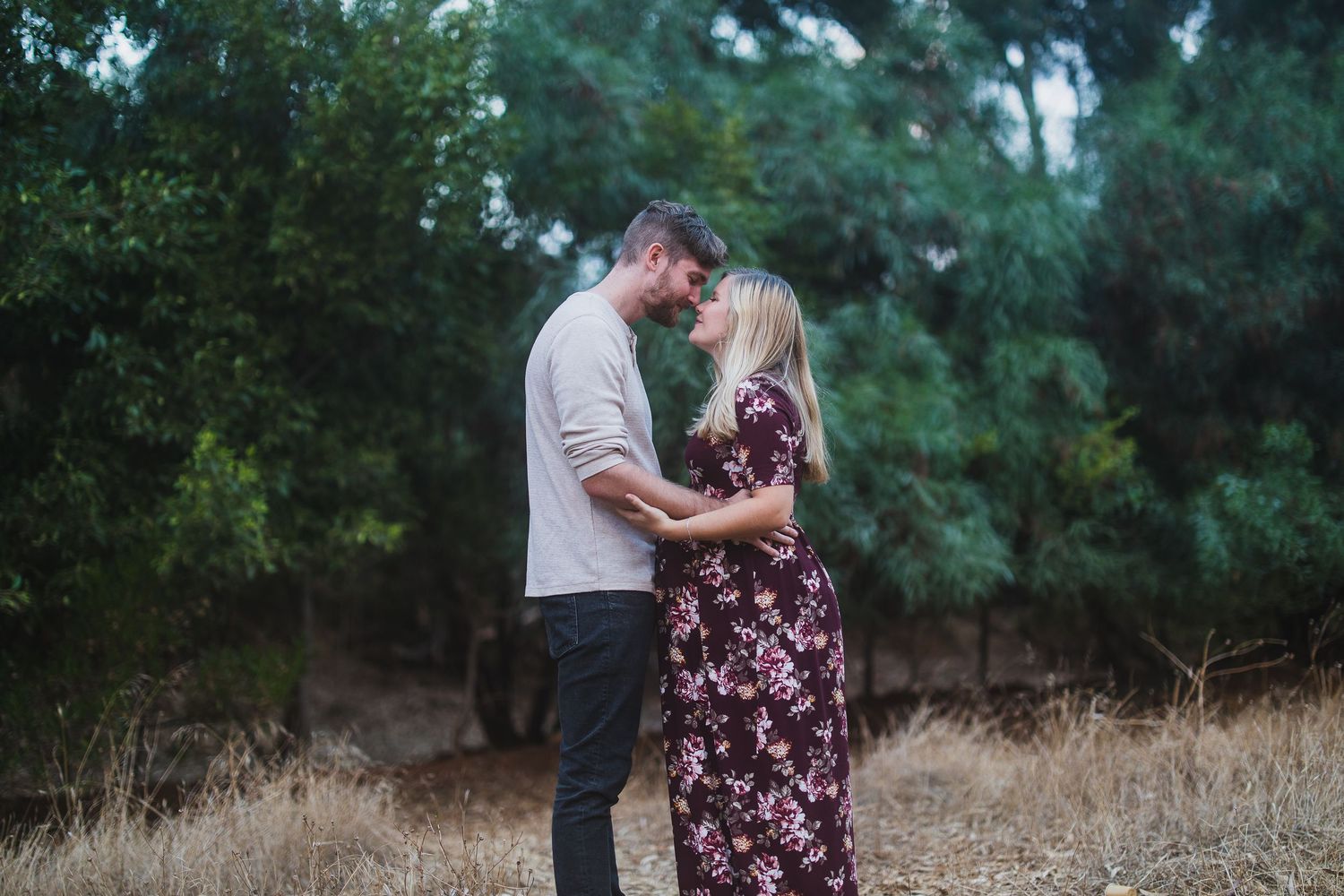 A couple shares an intimate moment outdoors wearing casual attire against a backdrop of green trees.