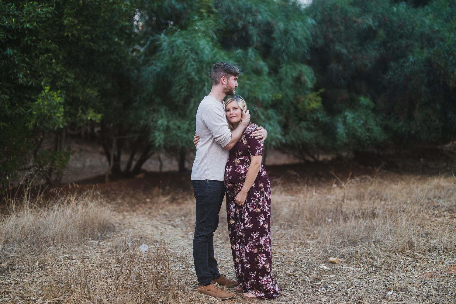 Couple embraces in an outdoor setting with dried grass and green trees behind them.