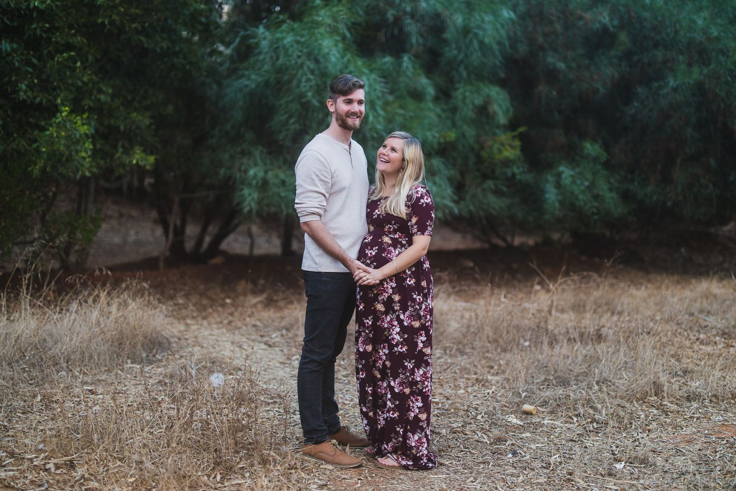 A loving couple embraces while standing in a field with evergreen trees in the background.