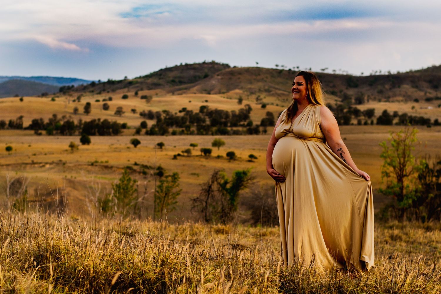 Silhouetted figure in flowing beige dress stands on hilltop against scenic rural landscape at golden hour.