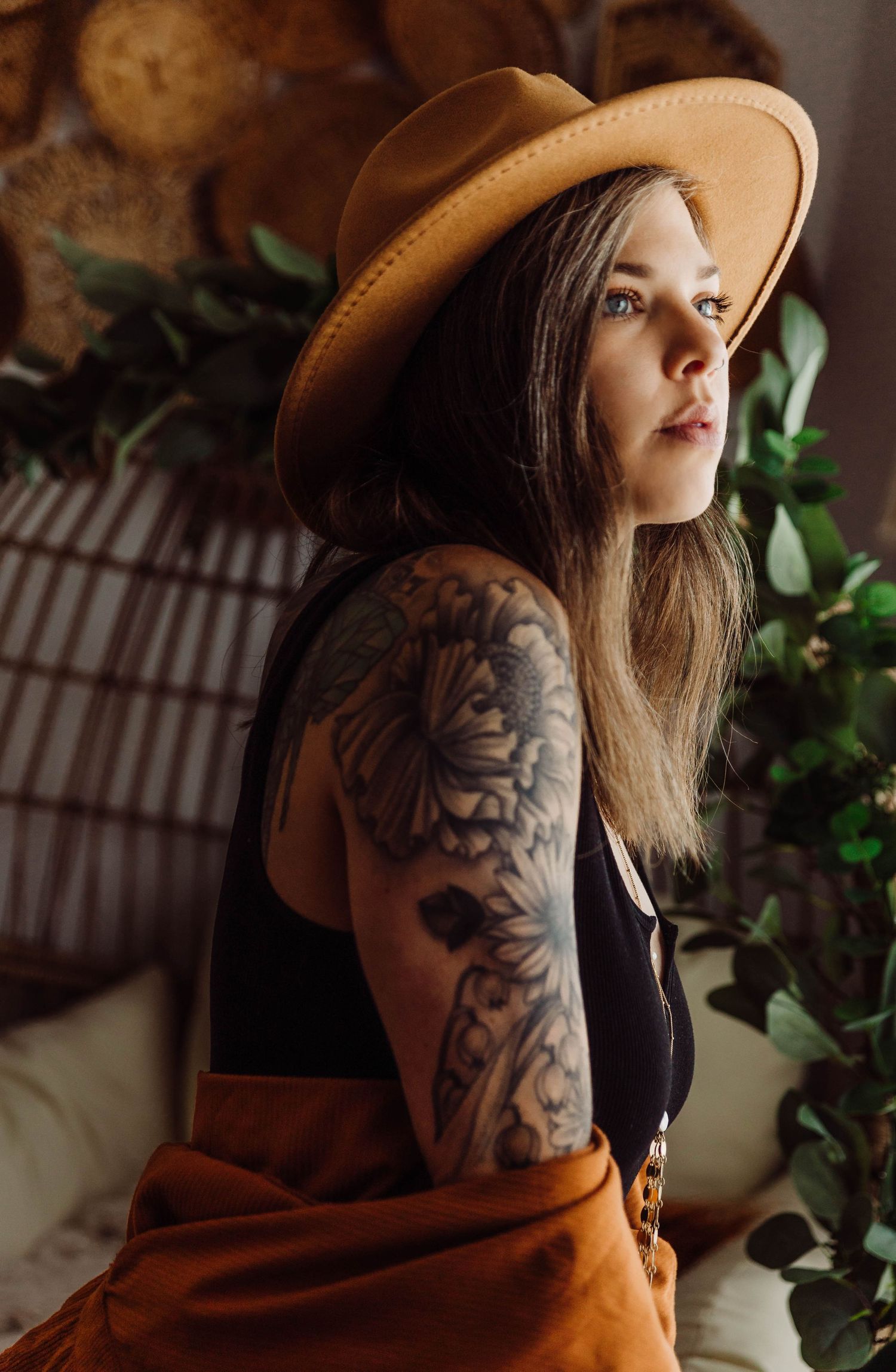 A woman in a brown hat with tattoos poses artistically against a natural backdrop.