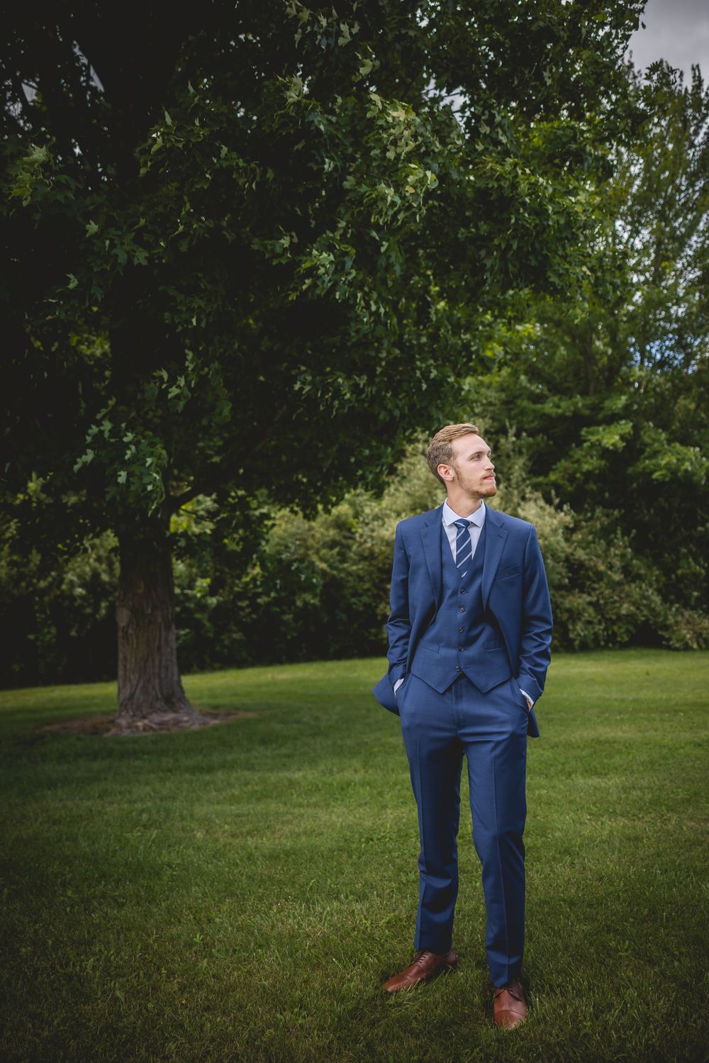 Man in blue suit poses thoughtfully on green lawn with tree in background.