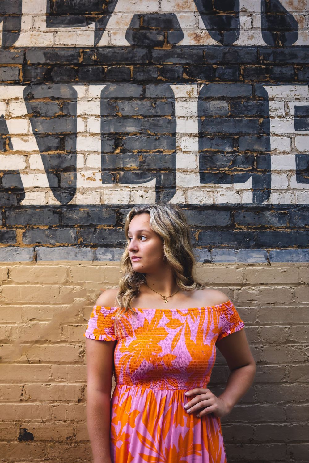 A summer portrait against a painted brick wall wearing a bright orange and pink floral dress.