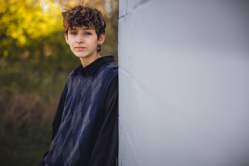 Person in navy hoodie leans against grey wall with autumn colors in background.
