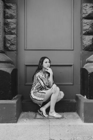 A woman in a striped dress sits contemplatively on stone steps against a gray door.