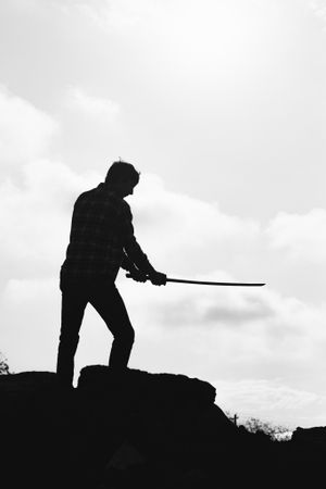 Silhouette of person holding staff while standing on rocky outcrop against cloudy sky.