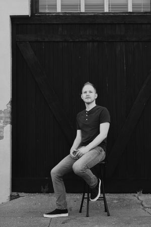 Monochrome portrait of person seated on stool against dark barn door background.