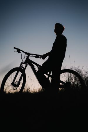 Silhouette of mountain biker standing with bicycle against sunset sky.