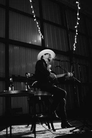 Black and white photo of a musician performing on stage with string lights in background.