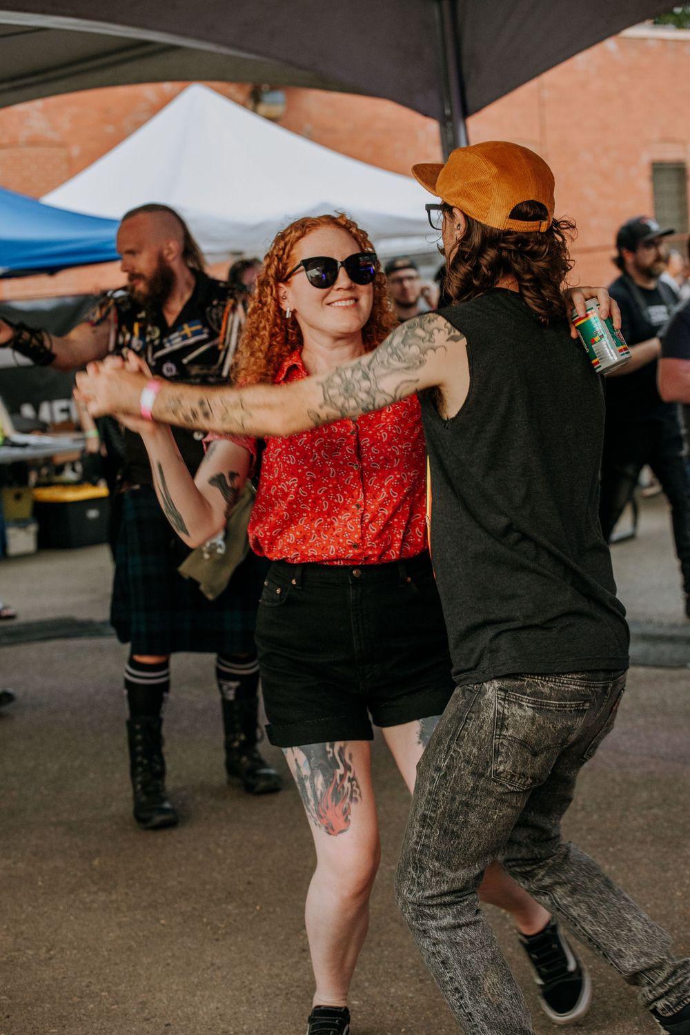 People dancing together at outdoor event with tents and vendors in background.