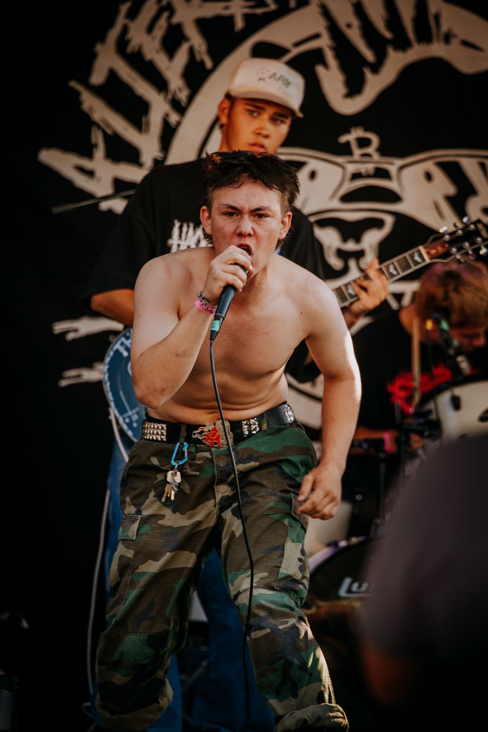 A shirtless performer in camo pants sings intensely on stage with band members behind.