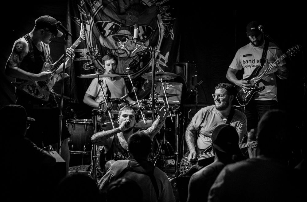 A black and white photo captures a high-energy band performance with multiple musicians on stage.