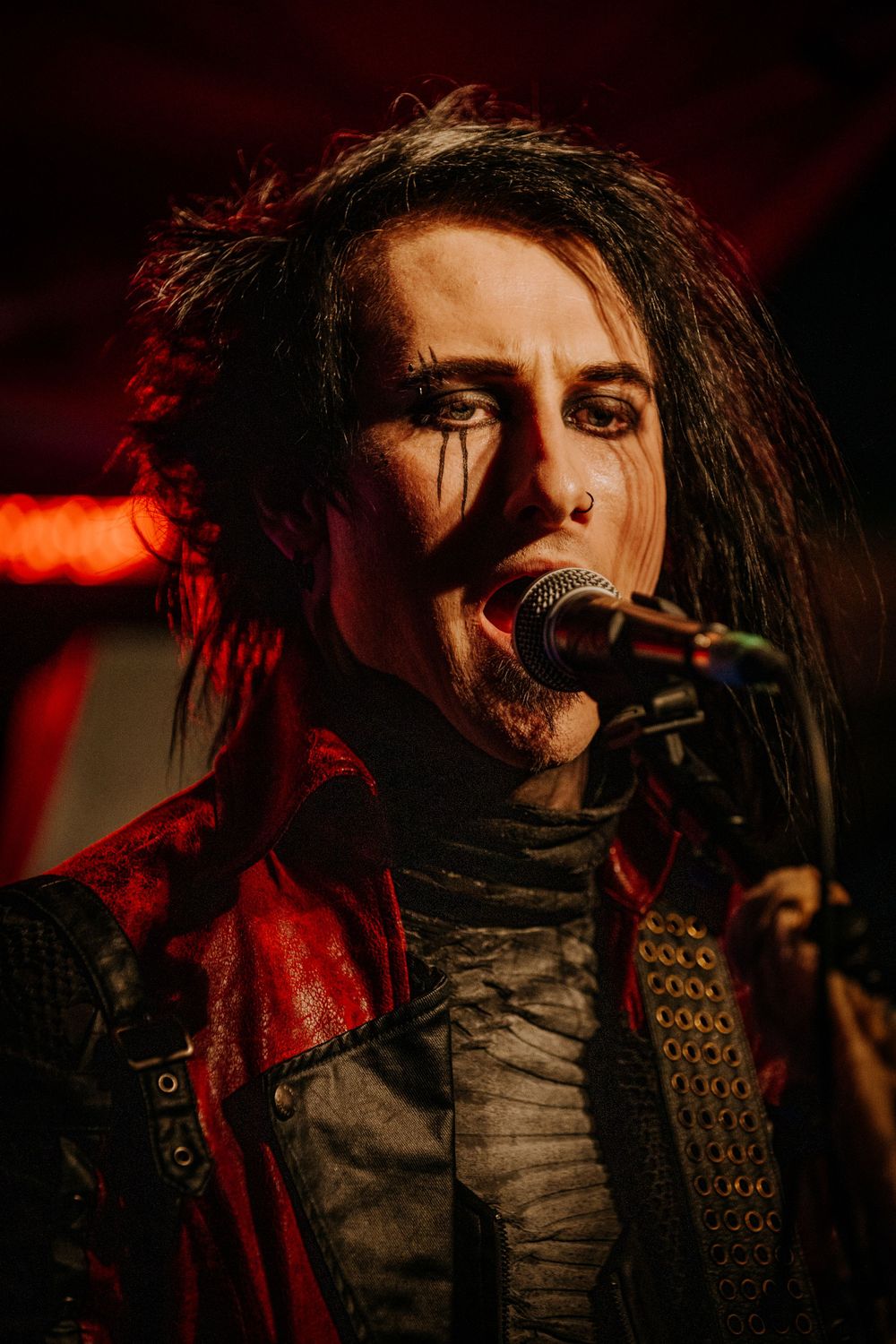 A dramatic performer in gothic attire and dark makeup sings into a microphone under red lighting.