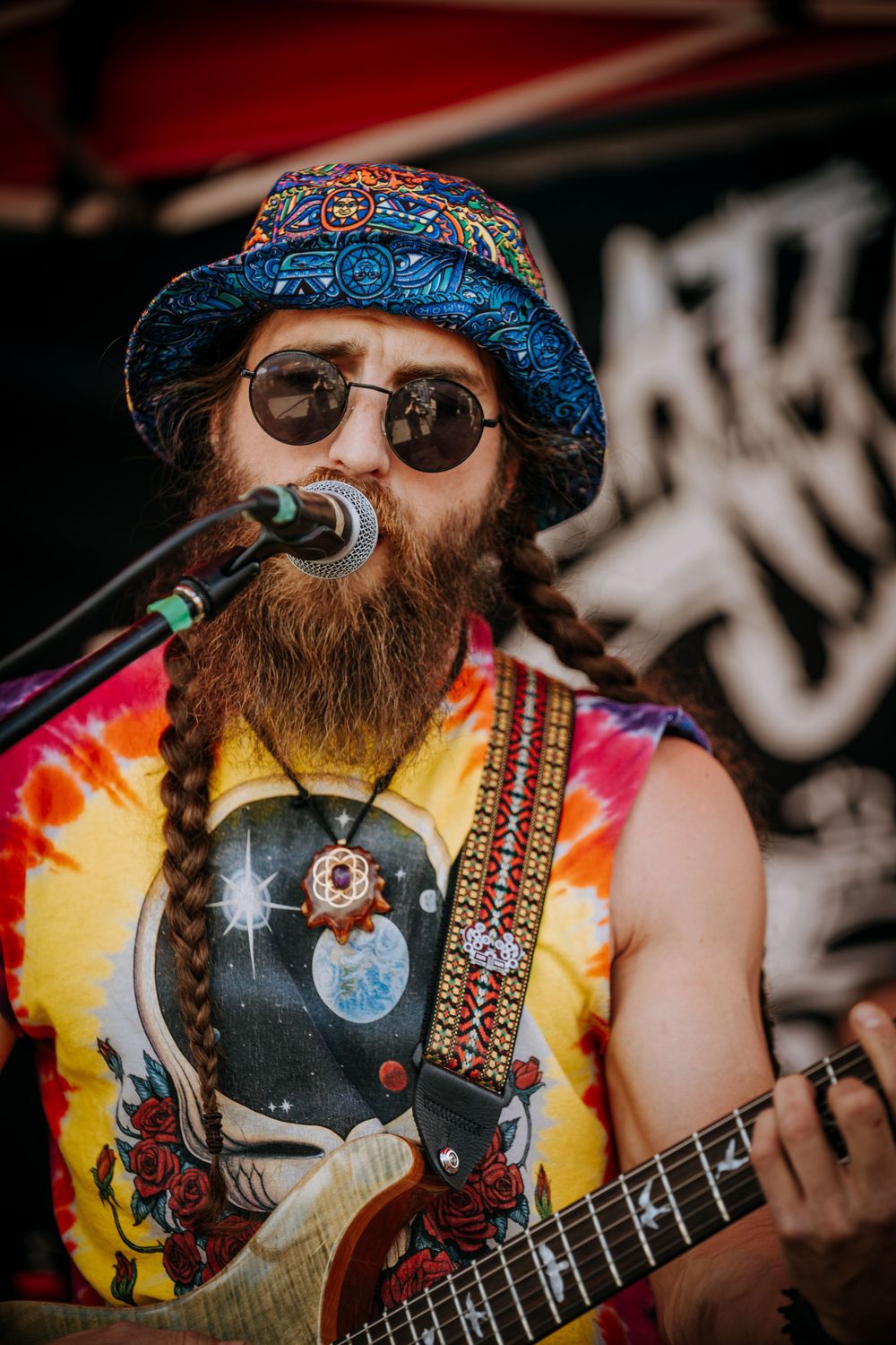 Musician in tie-dye tank top and patterned hat performs with guitar and microphone on stage.