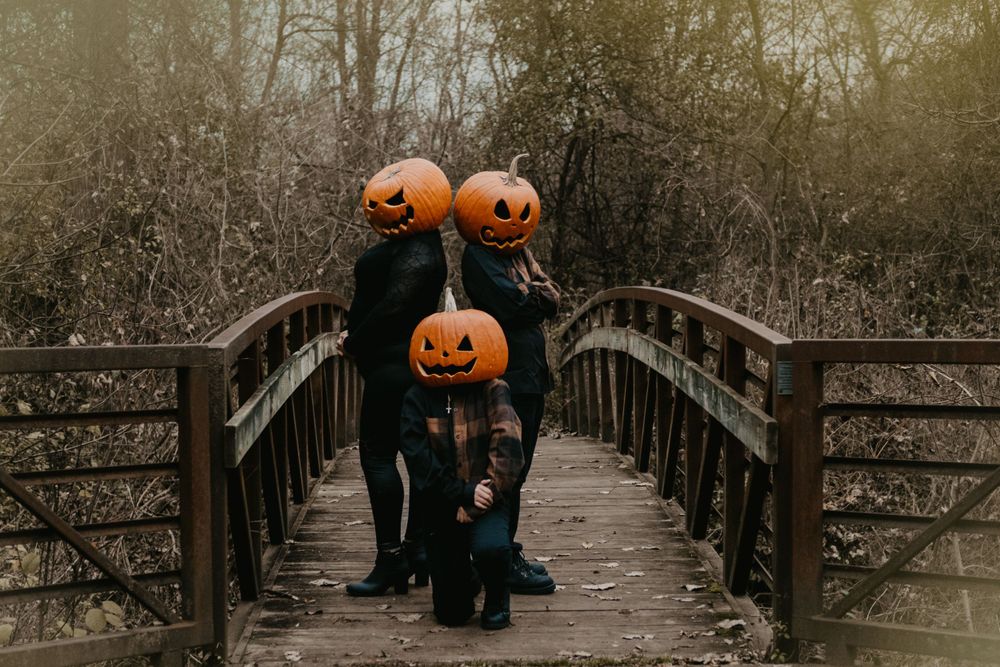 Three people with carved jack-o-lantern heads posing on wooden bridge surrounded by autumn trees.