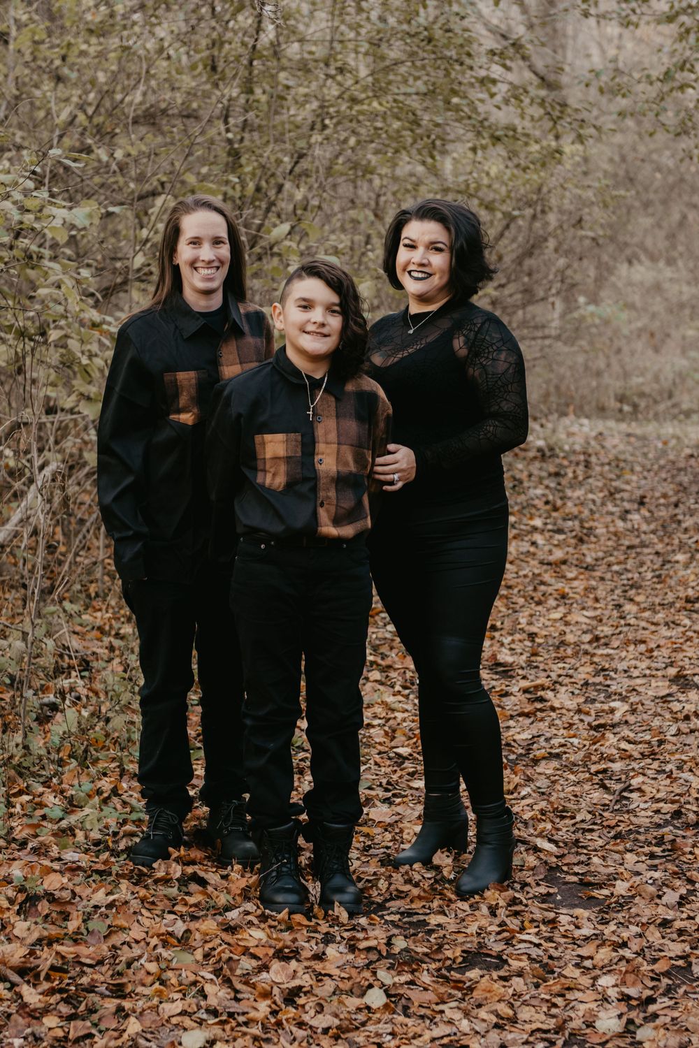 Three siblings posing on leaf-covered path in autumn woods, wearing coordinated black and plaid outfits.