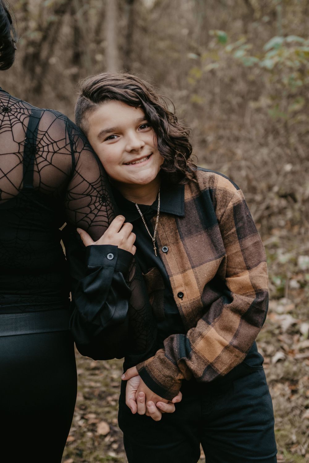 Young boy in plaid flannel shirt leaning against woman in black lace outfit in autumn forest.
