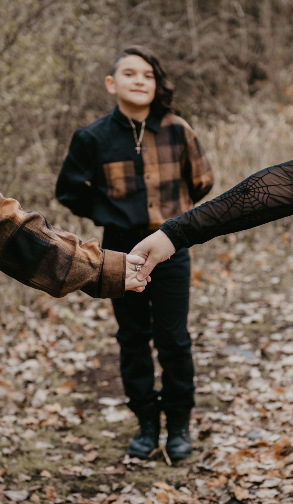 Boy in plaid shirt smiling while holding hands with two adults on a leaf-covered forest path.