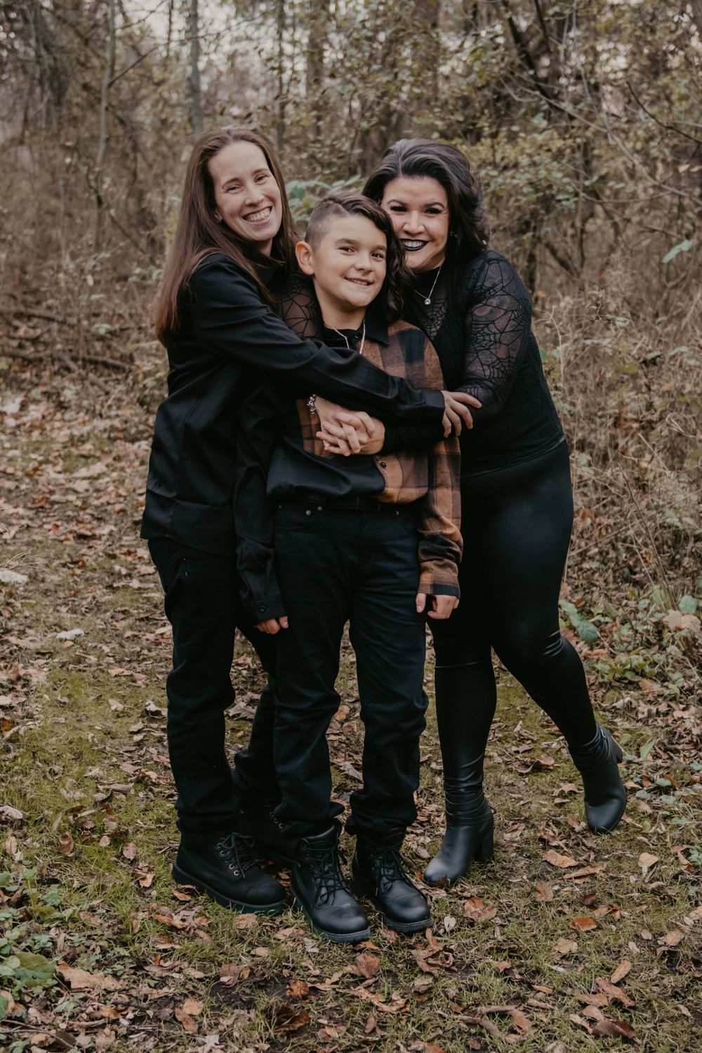 Two women hugging smiling boy in autumn forest, all wearing coordinated black and plaid outfits.