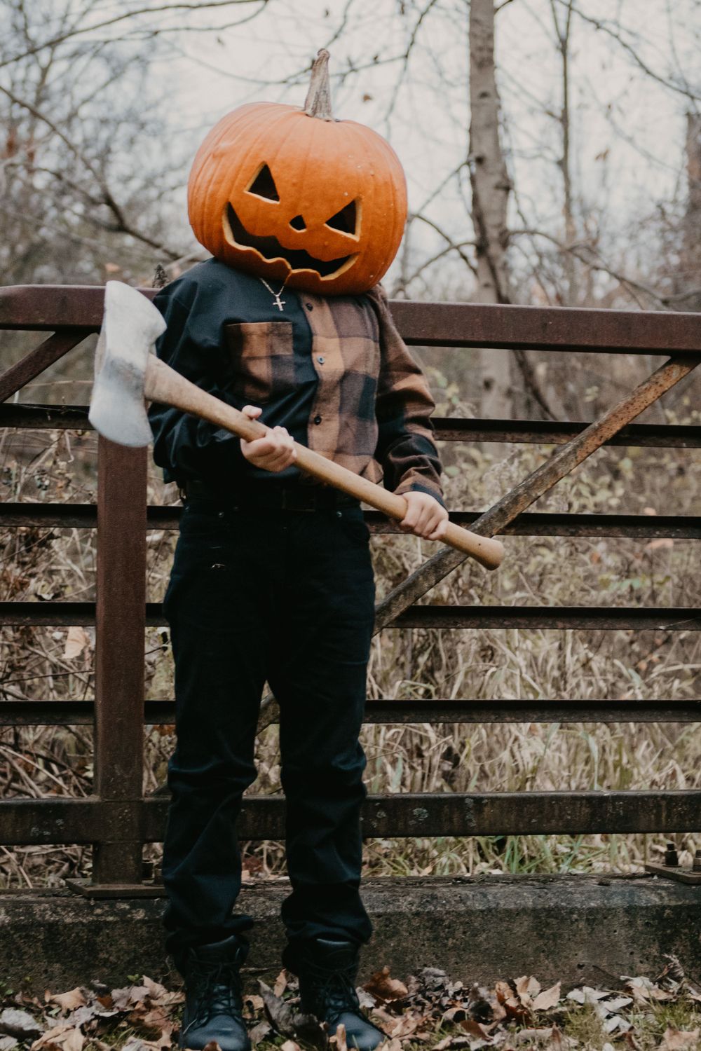 Person wearing carved jack-o-lantern head holding axe while standing near rustic wooden fence outdoors.