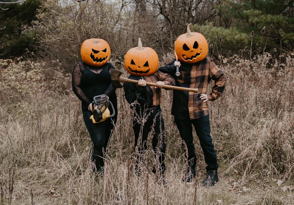 Three people holding axes wearing carved jack-o-lanterns on heads standing in dry autumn field.