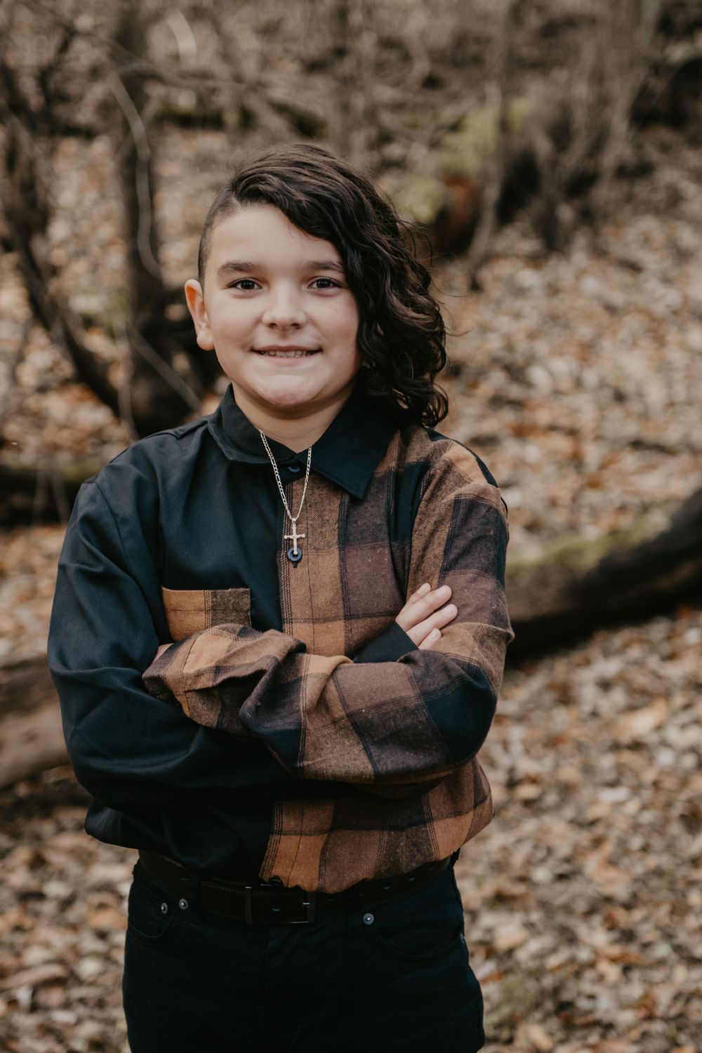 Smiling boy with curly hair in plaid flannel shirt, arms crossed, wearing cross necklace in autumn woods.