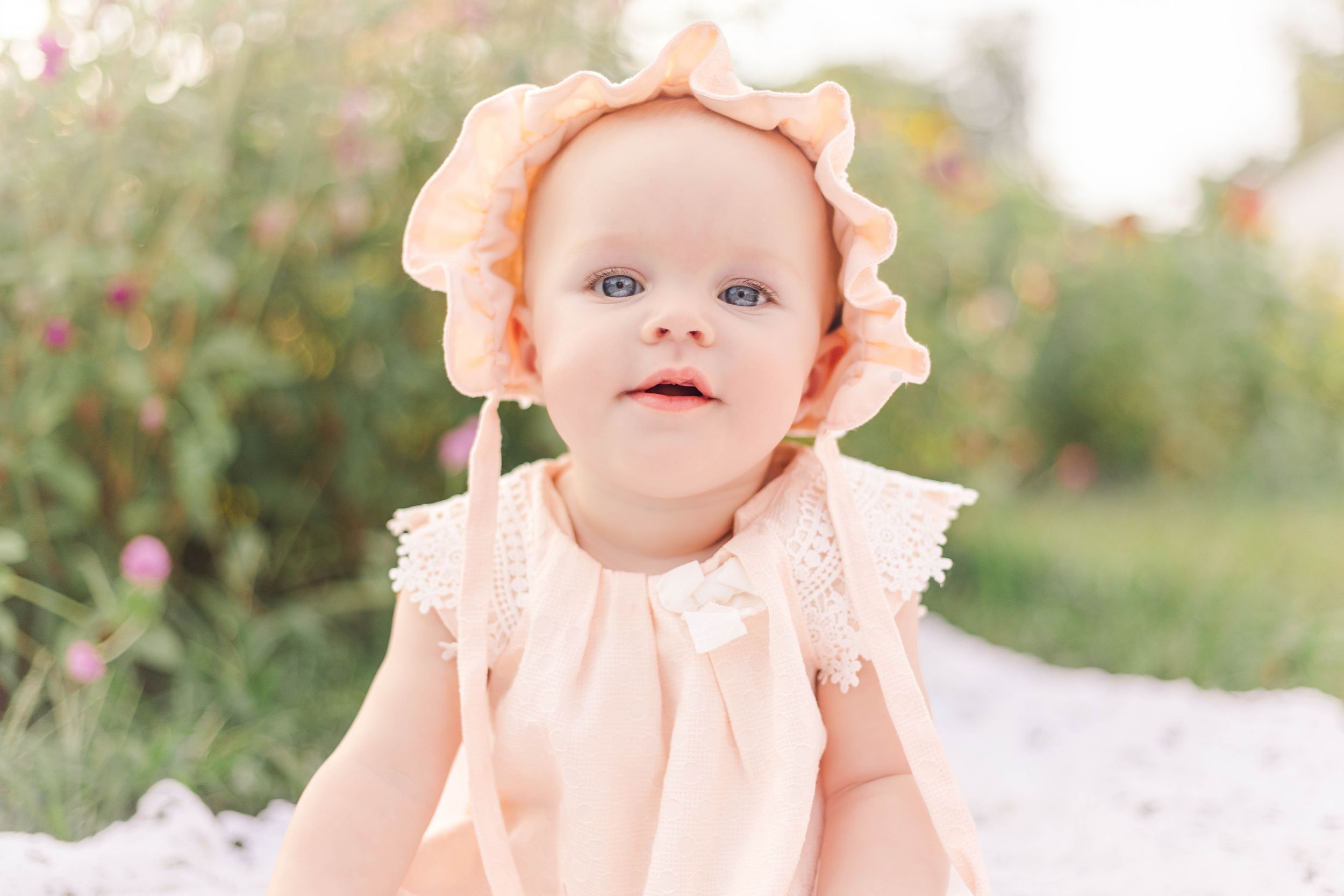 Baby girl wearing a peach bonnet and matching outfit sitting outdoors among flowers during a natural light milestone photography session.