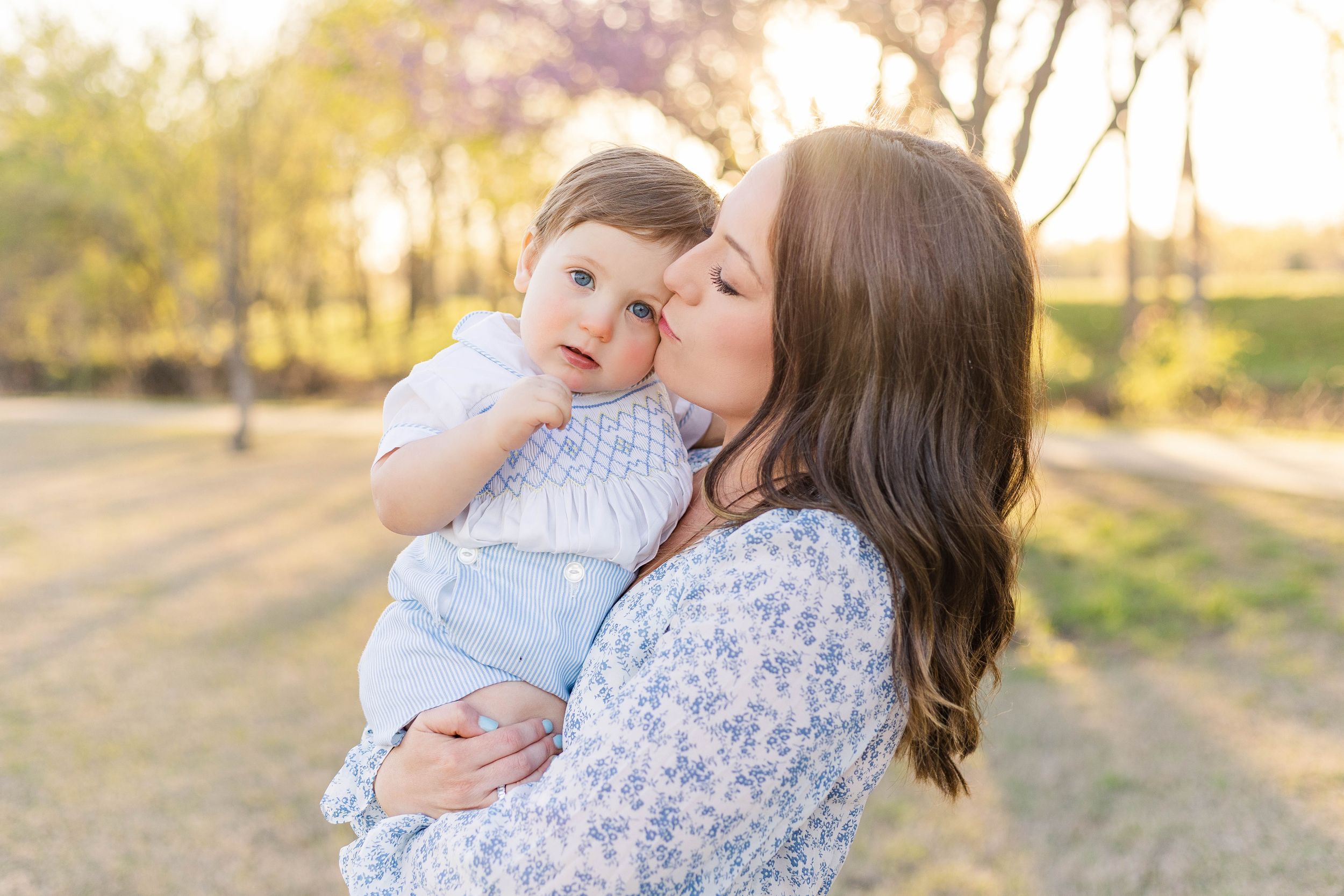 Mother holding her baby close and kissing their cheek outdoors during a golden hour motherhood photography session.