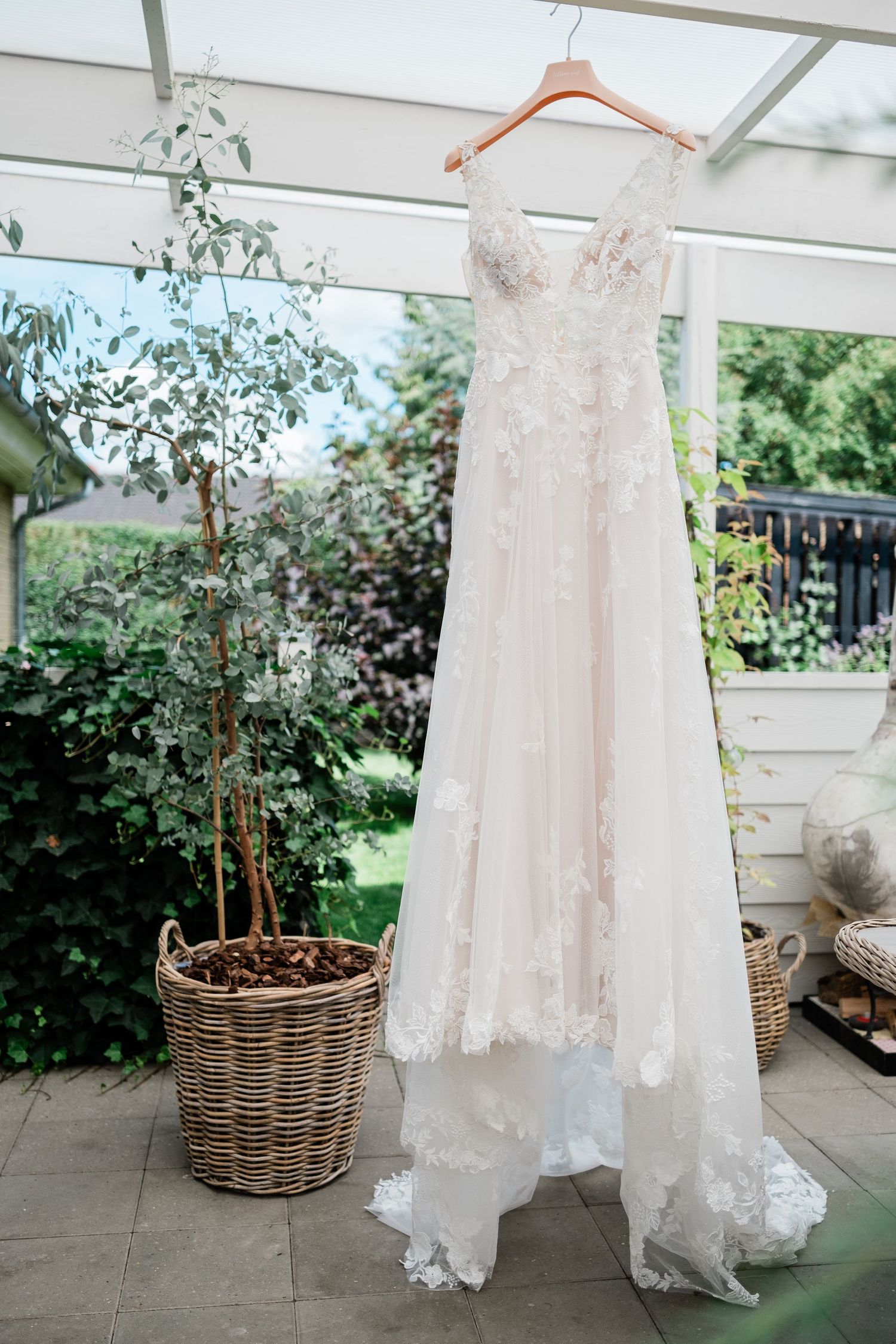 A delicate lace wedding dress hanging outdoors against greenery and wicker baskets.