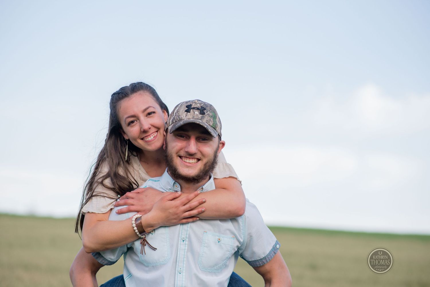 A couple shares a piggyback ride moment against a bright blue sky.