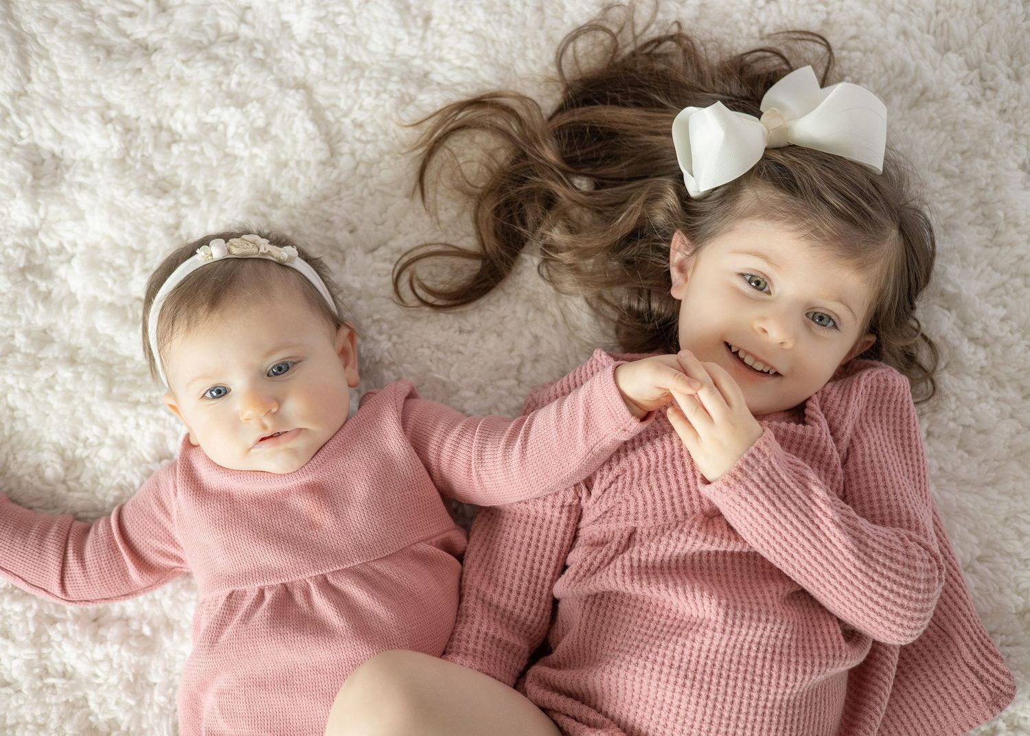 Two sisters wearing matching pink outfits and hairbows posing together on a white carpet