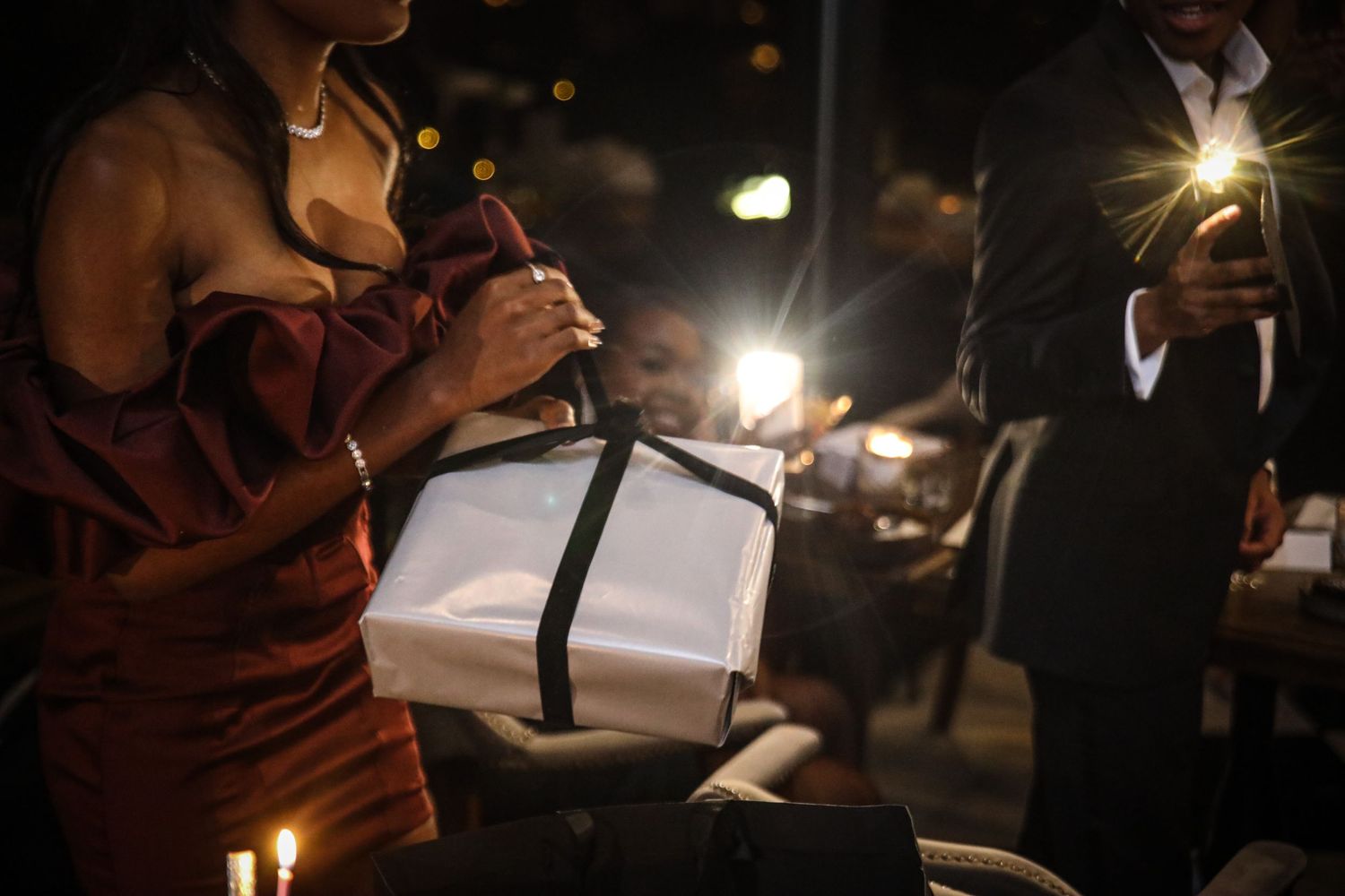 Guest in red dress unwrapping elegant white gift box at candlelit celebration.
