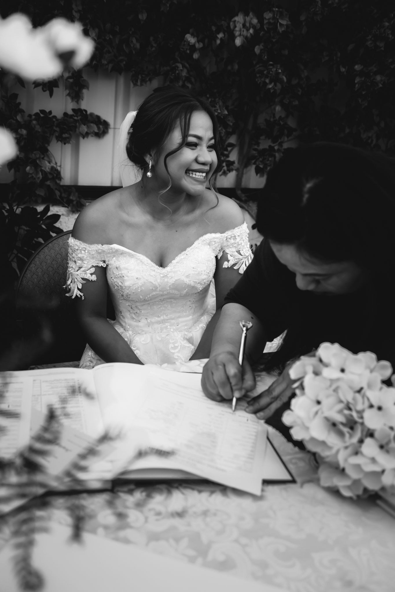 A couple signing marriage documents at a table with a bouquet of flowers.