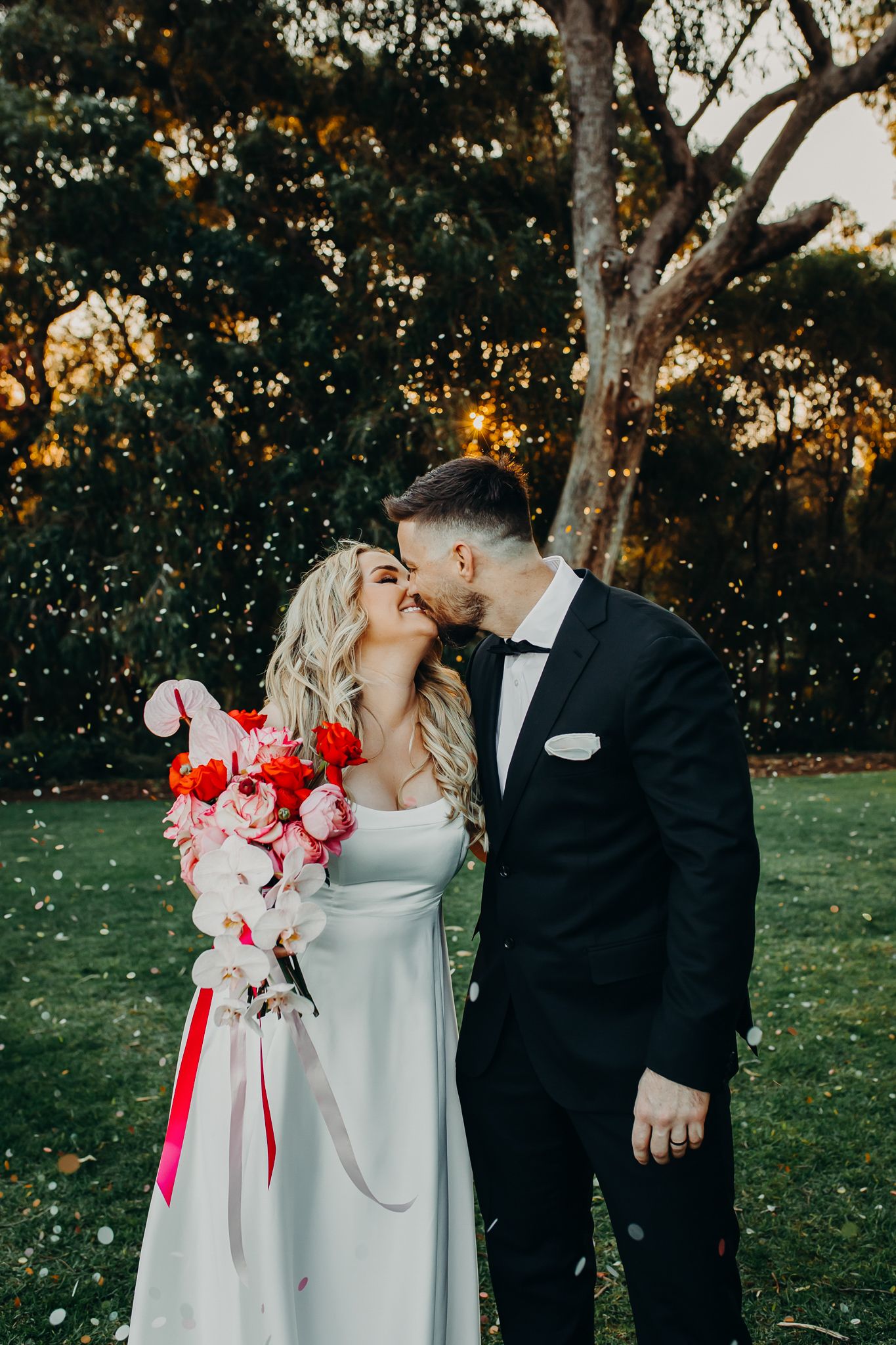 A wedding couple shares a romantic kiss in a garden setting at sunset with a red and pink floral bouquet.