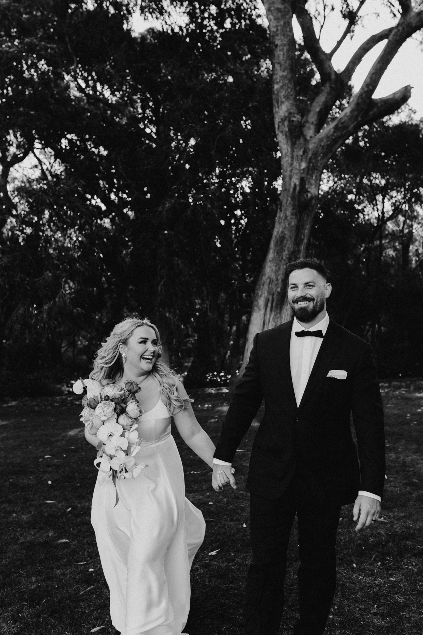 Black and white portrait of a laughing wedding couple walking outdoors among trees.
