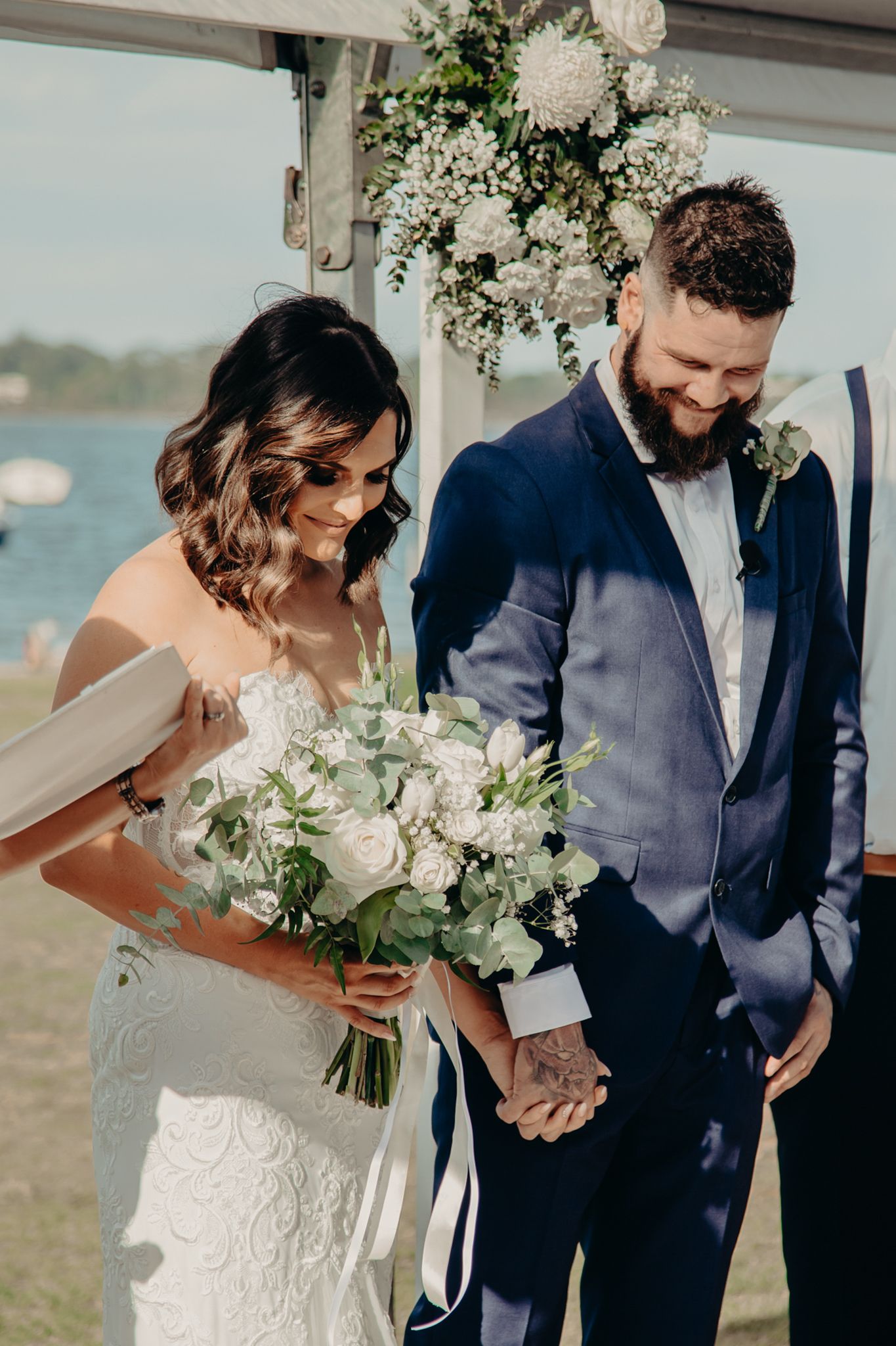 Newlyweds stand under floral arch by waterfront during outdoor ceremony.