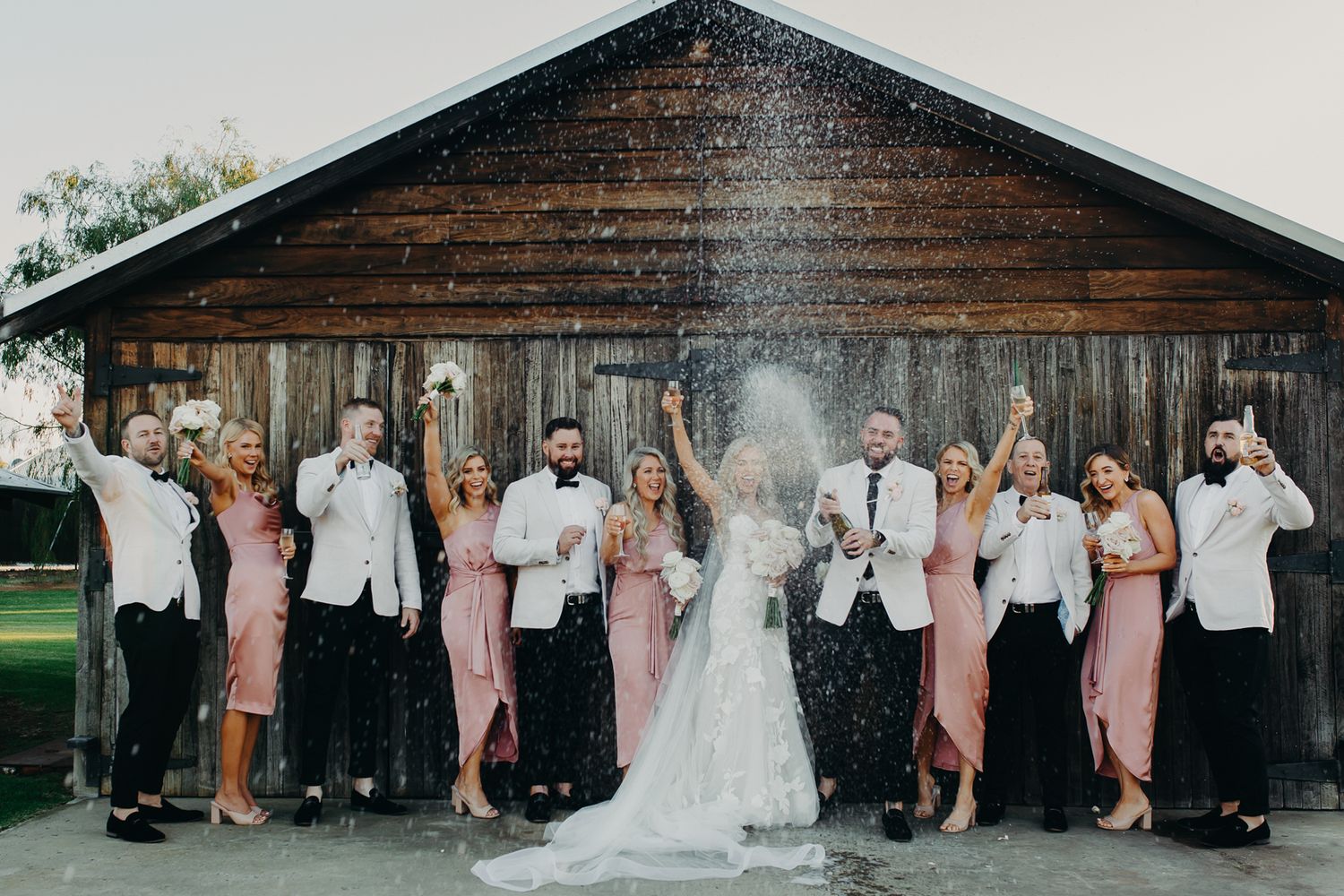 Wedding party pops champagne in front of rustic barn backdrop.