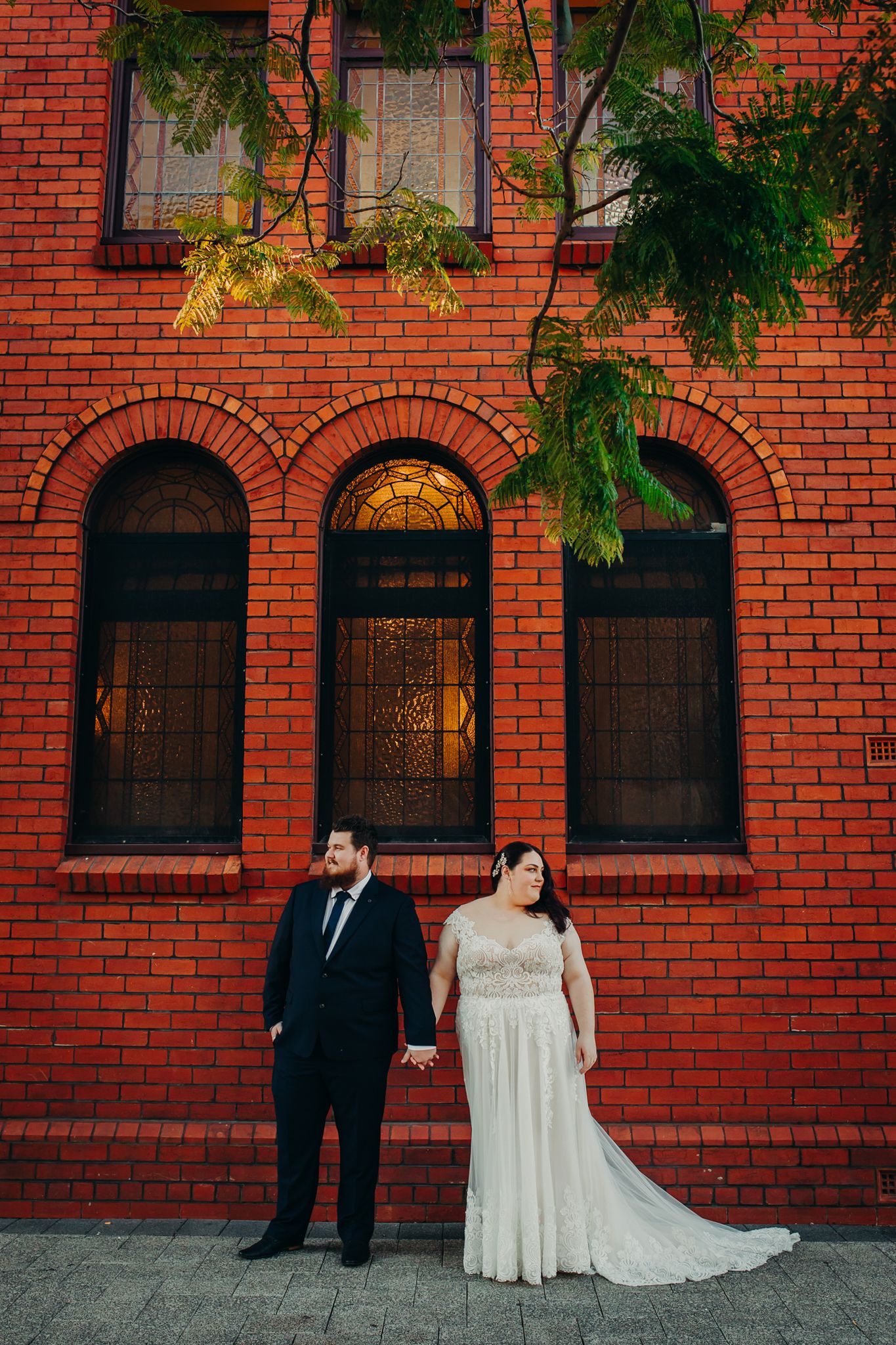 Wedding couple poses against a classic red brick building with arched windows.