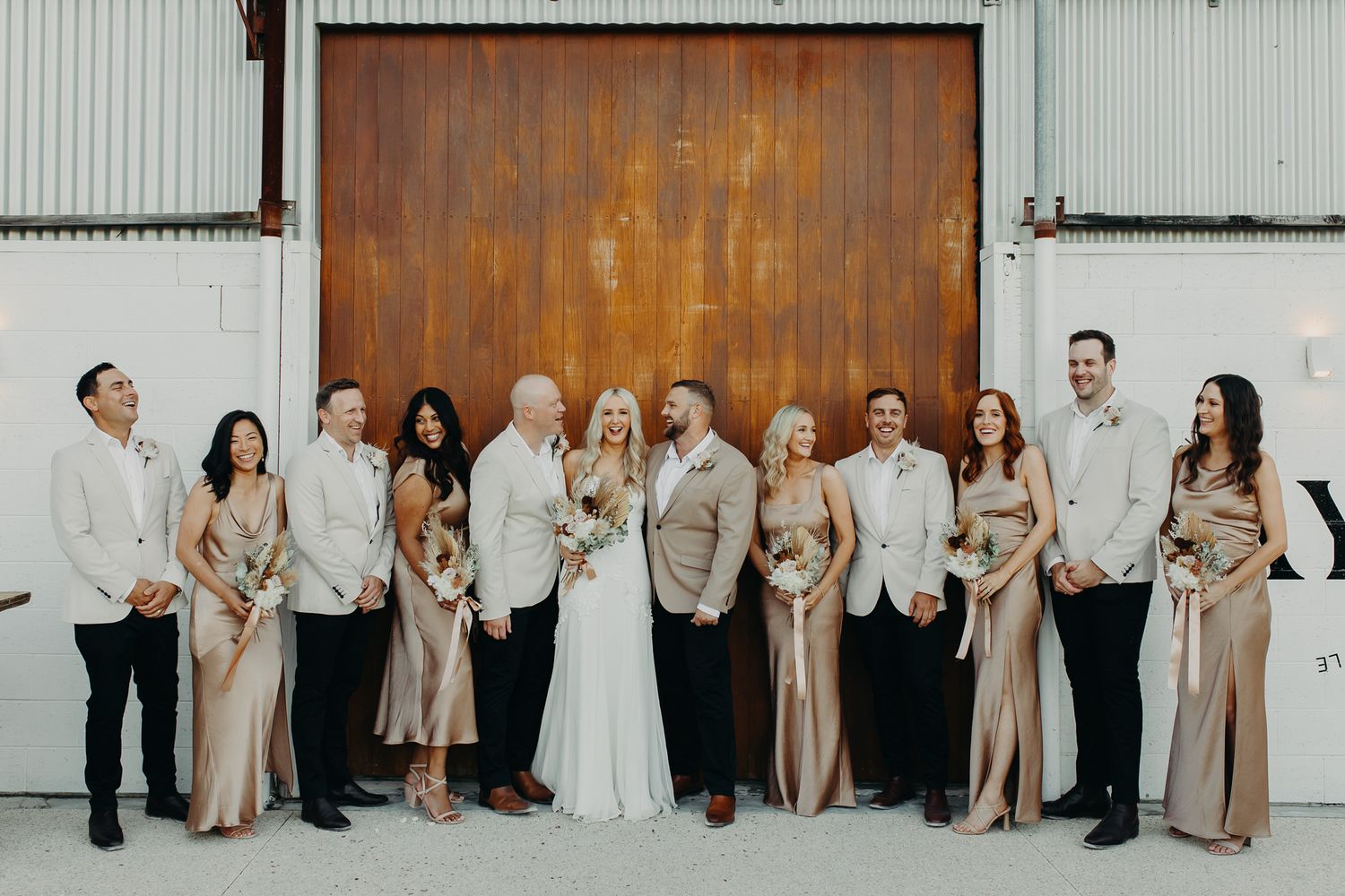 Wedding party in neutral tones poses formally against white industrial wall.