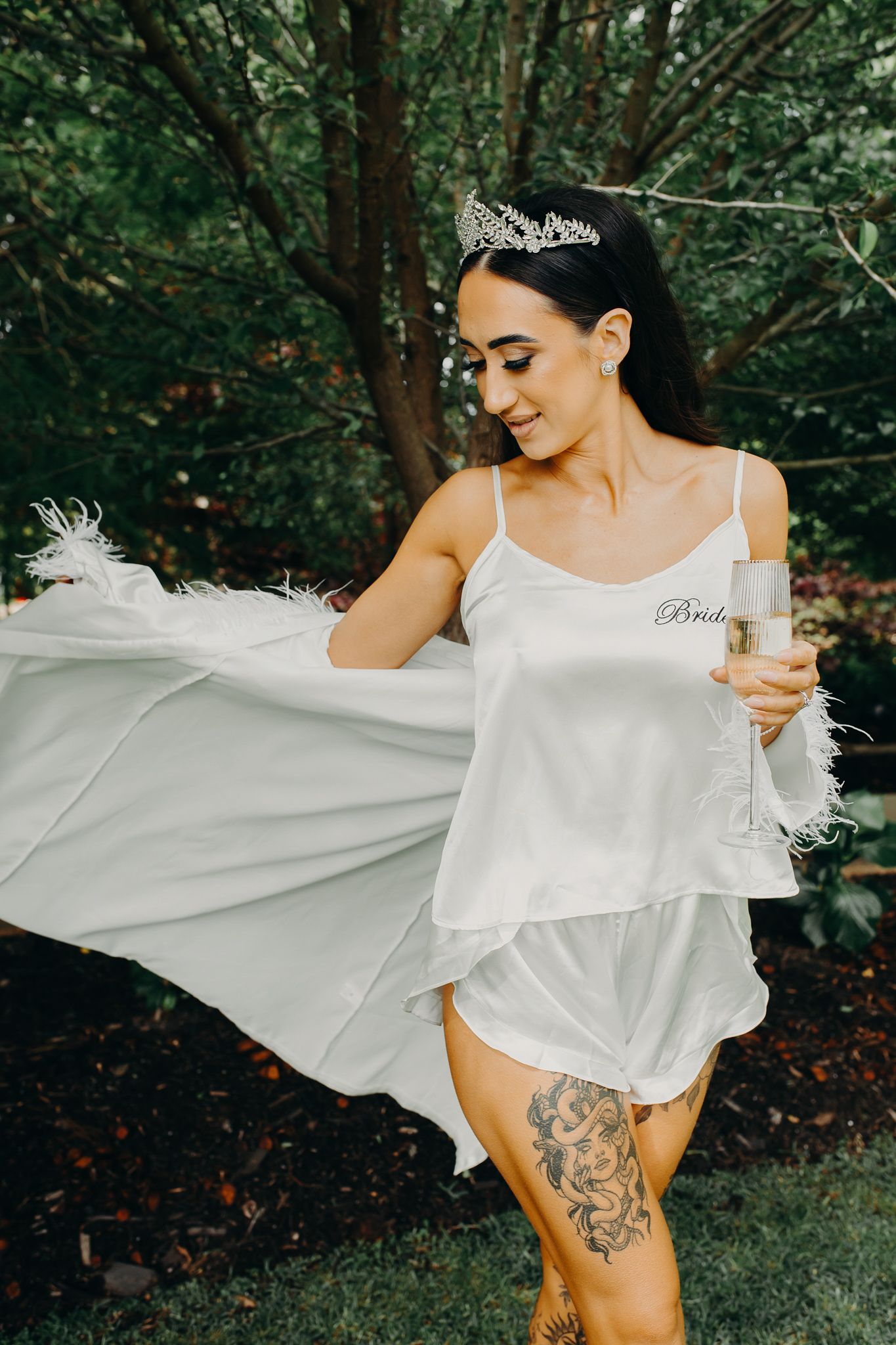 Bride in white silk loungewear twirls with champagne in forest setting.